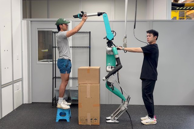 A researcher stands on a small stool to reach the raised neck-mounted SRL, with another researcher beside the humanoid.